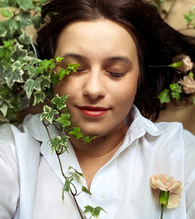 Dominika Bęben w białej koszuli, z zamkniętymi oczami, wśród gałązek bluszczu i kwiatów.