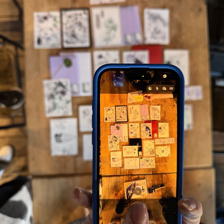Dłonie trzymające telefon, który fotografuje ułożone na stole kartki z odbitkami roślin.